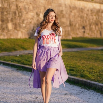 Mulher com t-shirt branca 'EXOTIC' e saia lilás a caminhar em calçada de pedra