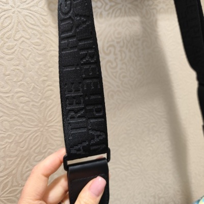 Correia preta com padrão de letras em relevo e detalhes em couro segurada por mão com unhas rosas.