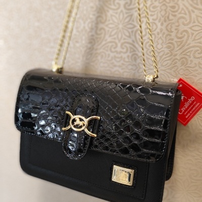 Mala preta com textura pele de cobra e alça dourada pendurada numa parede beige