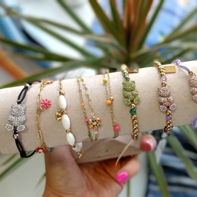 Várias pulseiras coloridas em suporte bege, com detalhes florais e contas, seguradas numa mão em ambiente ao ar livre
