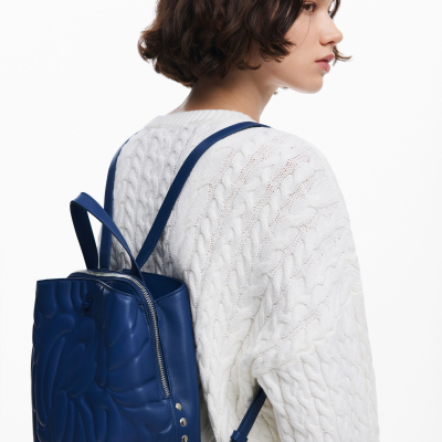 Mochila azul em pele com padrão em relevo e fecho prateado, usada por pessoa com camisola branca de malha