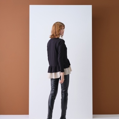 Mulher de costas com casaco preto e bainha cinza, calças de couro e botas de salto, em fundo castanho com painel branco