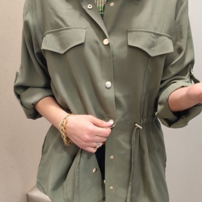 Mulher a usar casaco verde-oliva com botões e cordão na cintura, pulseira dourada, fundo interior simples.