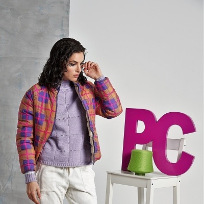 Mulher com casaco xadrez rosa e lilás e camisola lilás ao lado de cadeira branca com letras 'PC' cor de rosa e lâmpada verde