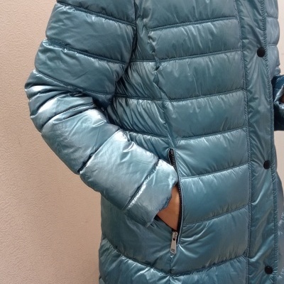 Casaco acolchoado azul esverdeado com fecho e botões em fundo neutro.