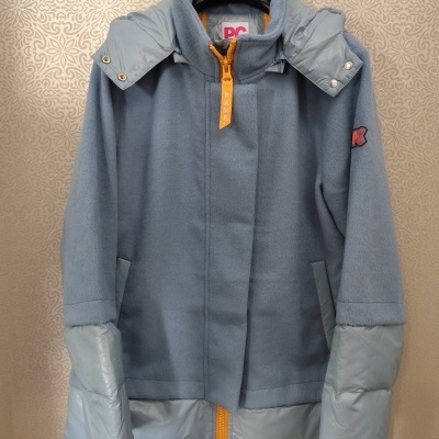 Casaco azul claro com mistura de tecidos e fecho amarelo pendurado numa parede com padrão abstrato