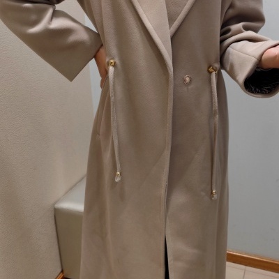 Casaco comprido bege claro com gola de lapela e detalhe de cordões dourados numa sala interior