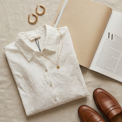 Camisa branca bordada, colar dourado, brincos de argola, sapatos castanhos e livro aberto.