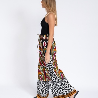 Mulher vestindo calças largas estampadas multicoloridas, top preto com alças de contas bege e sandálias pretas, vista lateral em fundo branco.