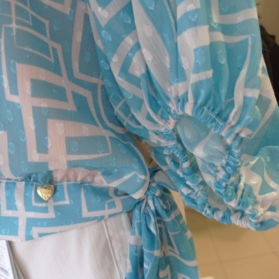 Blusa azul com padrão geométrico branco e mangas bufantes com laço e pendente dourado