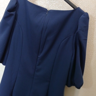 Vestido azul escuro com mangas frisadas pendurado num cabide preto numa parede clara com padrão decorativo