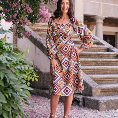 mulher com vestido midi geométrico colorido, escada de pedra e flores roxas ao fundo