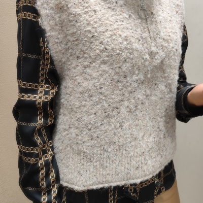 Colete cinza claro felpudo com fecho de correr e gola alta, sobre camisa preta com padrão geométrico dourado e calças bege.