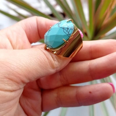 Anel dourado com pedra turquesa e veios pretos numa mão com unhas rosa