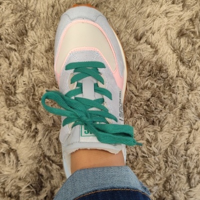 Sapatilha desportiva branca com detalhes rosa e cinza, atacadores verdes, sola laranja, em pé sobre tapete bege