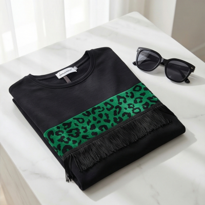 T-shirt preta com faixa verde estampada e franjas, com óculos escuros pretos ao lado sobre superfície branca