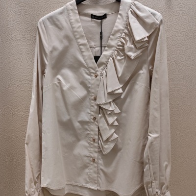 Blusa creme com botões dourados e folhos decorativos pendurada num cabide preto.