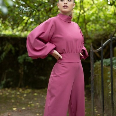 Mulher com macacão rosa de mangas bufantes em jardim com árvores e portão.