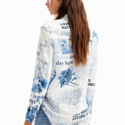 Mulher de costas a usar camisa branca estampada a azul com textos e desenhos, com calças azuis.