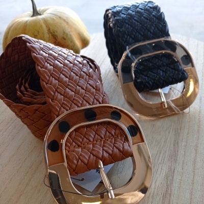 Dois cintos trançados, castanho e preto, com fivelas douradas grandes, sobre mesa de madeira e abóbora ao fundo