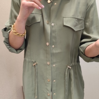 Parka verde oliva com botões e bolsos no peito, usada por pessoa com joias e camisola às riscas.
