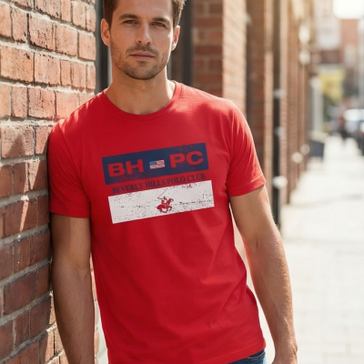 Homem com t-shirt vermelha com estampa Beverly Hills Polo Club encostado em parede de tijolos em rua urbana