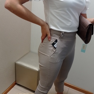 Pessoa com blusa branca de mangas bufantes, calças cinza claras justas e sapatos nude com carteira castanha num interior.