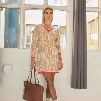 Mulher vestindo vestido bege com letras e bordas vermelhas, meias bege, sapatos pretos, segurando mala castanha em ambiente interior