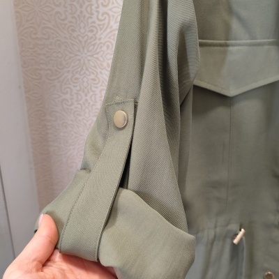 Casaco verde-oliva com botão metálico e cordão ajustável, visto em fundo com padrão decorativo