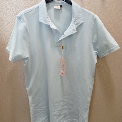 Polo azul claro de algodão com etiqueta pendurada e gola com botões numa parede decorada