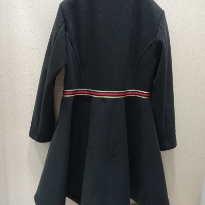 Casaco preto evasê com cinto riscado pendurado numa parede clara