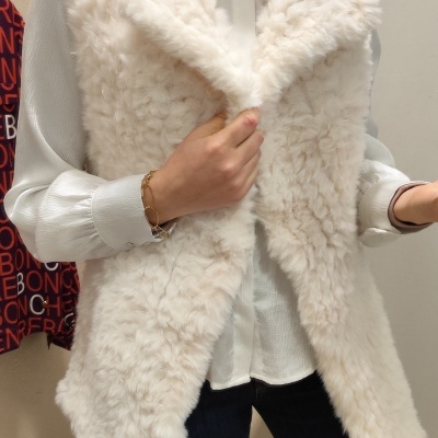 Colete feminino de pelo sintético branco com camisa branca e jeans escuro em ambiente interior