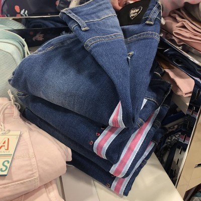 Calças de ganga azuis com riscas brancas e cor-de-rosa na bainha, empilhadas sobre superfície branca com outras roupas próximas