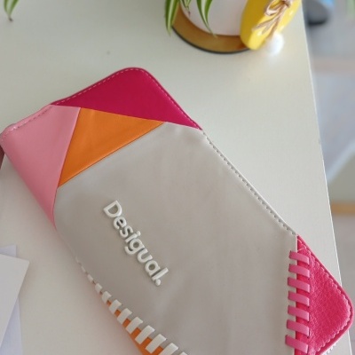 Carteira Desigual colorida em tecido sintético sobre mesa branca com planta ao fundo