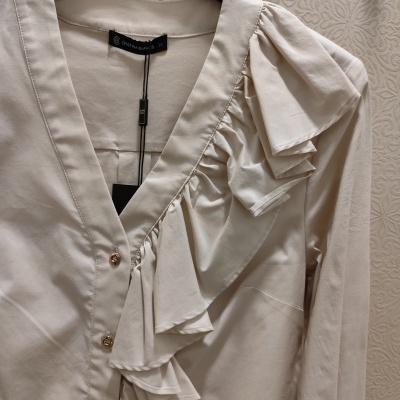 Blusa creme com folhos e botões dourados pendurada num cabide preto