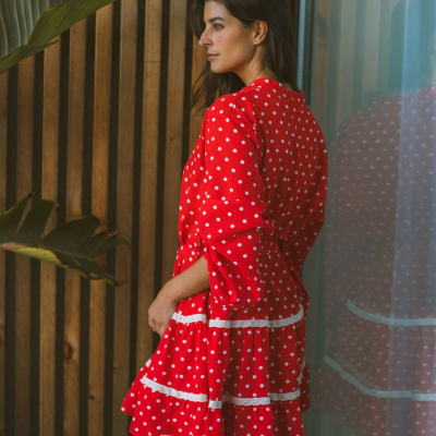 Vestido vermelho às bolinhas brancas com renda, usado por mulher em cenário com parede de madeira e planta