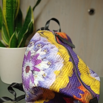 Mochila de tecido floral colorido com fecho preto ao lado de planta em vaso branco