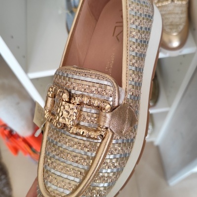 Sapato mocassim dourado com fivela decorativa e riscado bege segurado por mão num ambiente de loja