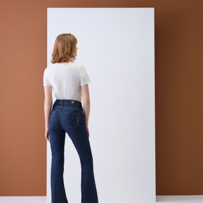 Mulher de costas com calças de ganga azul escuro, t-shirt branca e sapatos pretos em fundo branco e parede castanha.