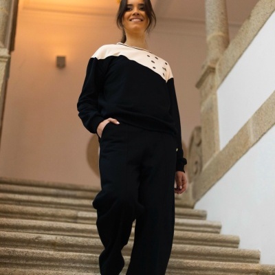 Mulher vestindo camisola preta e branca com calças pretas e sapatilhas brancas numa escadaria interior.