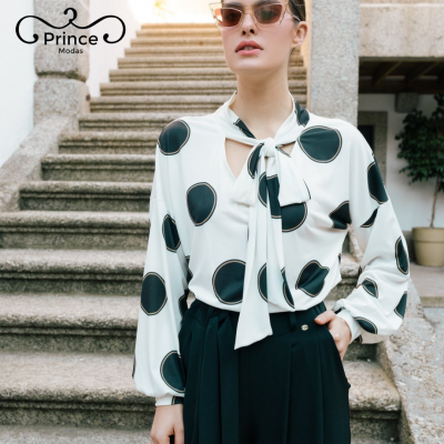 Mulher com blusa branca com pontos pretos e calças pretas junto a escadas de pedra