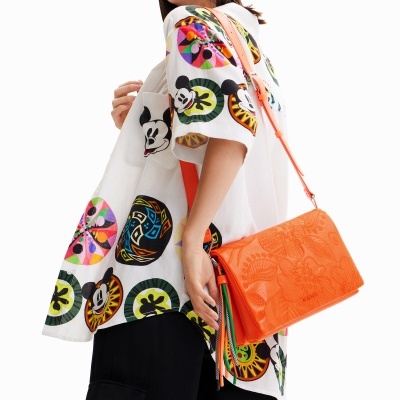 Mulher com camisa branca estampada colorida de Mickey Mouse, calças pretas largas e mala laranja ao ombro