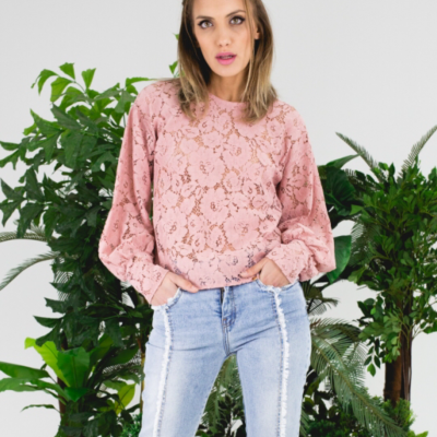 Mulher com blusa rosa de renda e calças de ganga azuis claras, plantas verdes ao fundo.