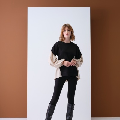 Mulher de pé com camisola preta e beje, leggins e botas pretas, parede castanha e painel branco