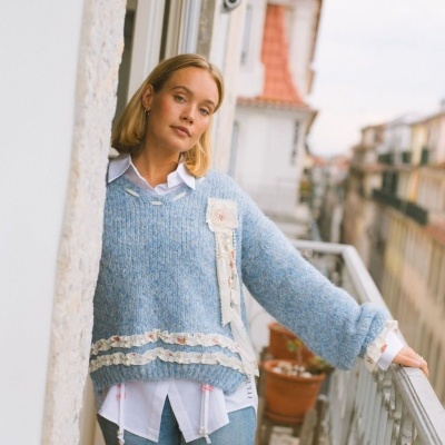 Mulher com camisola azul de malha com detalhes floridos e camisa branca numa varanda