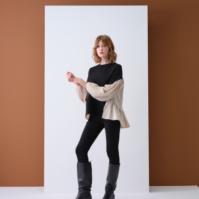 Mulher com blusa preta e beje, calças pretas e botas pretas em estúdio minimalista.