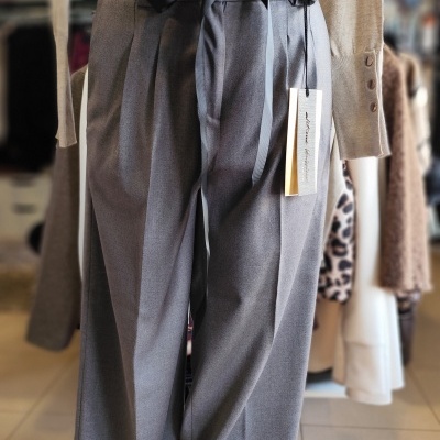 Calças cinzentas de cintura alta com laço preto em manequim, com suéter bege e loja ao fundo.