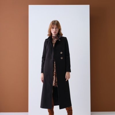 Mulher com casaco preto comprido, vestido animal print e botas castanhas, fundo branco e castanho