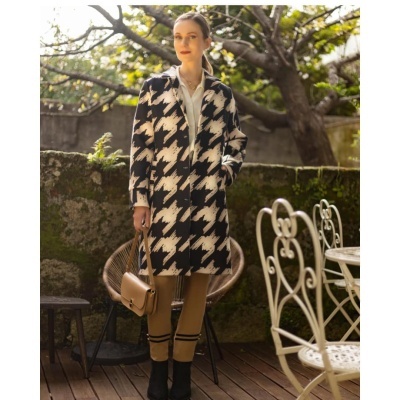 Mulher com casaco pied-de-poule preto e branco, calças camel, mala castanha e ténis pretos num jardim exterior