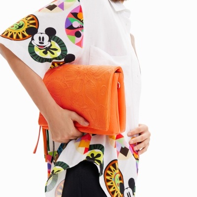 Pessoa de perfil com camisa com padrão do Mickey Mouse e mala laranja texturizada.